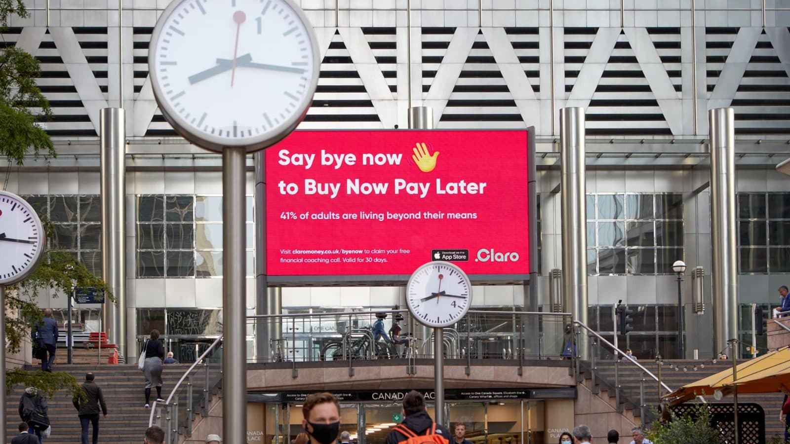 A red Claro billboard reading "Say bye now to Buy Now Pay Later" above the steps at Canada Place, Canary Wharf, with commuters passing