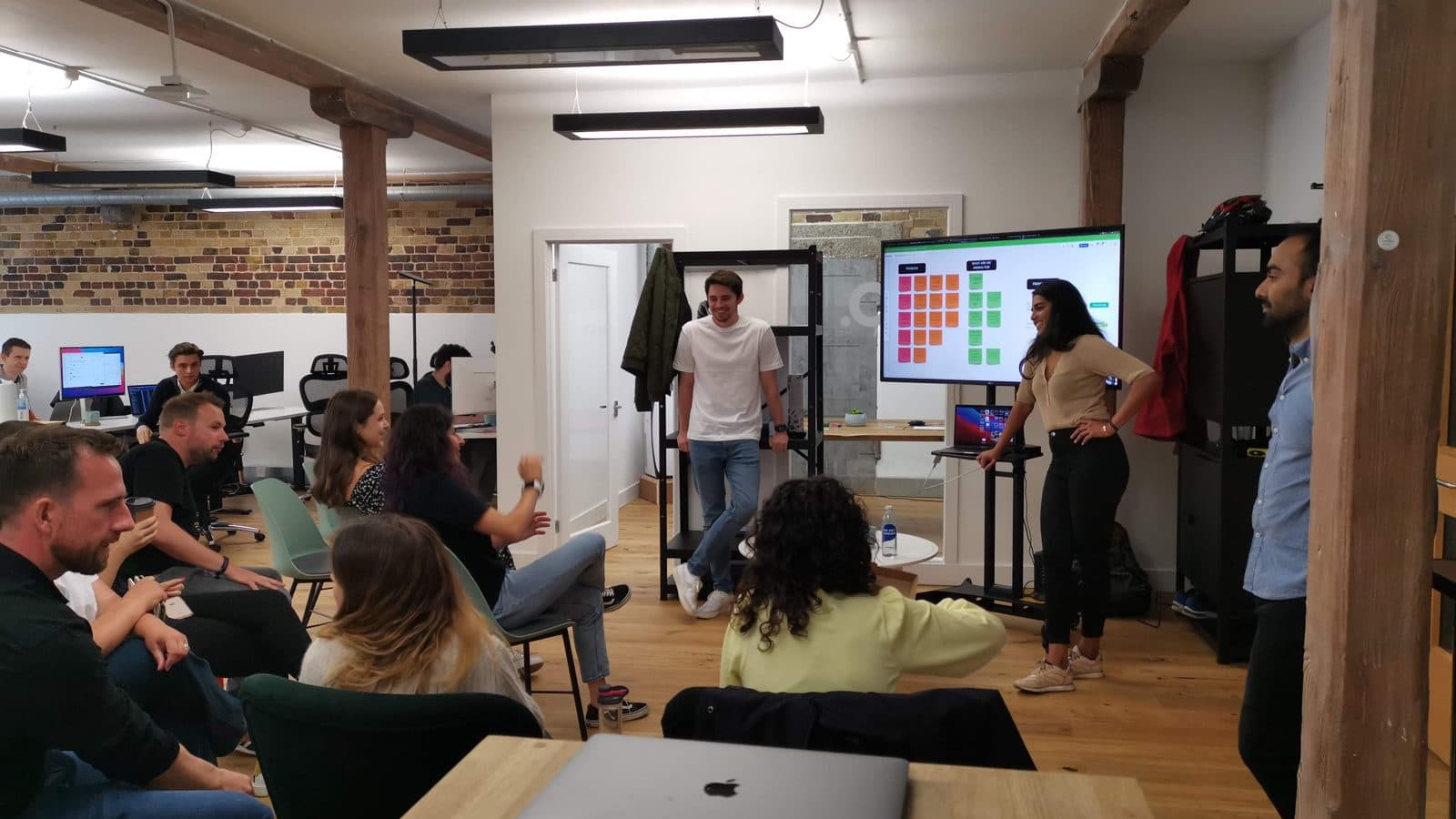 A team gathered in a brick-walled office, watching a colleague present at a wall screen